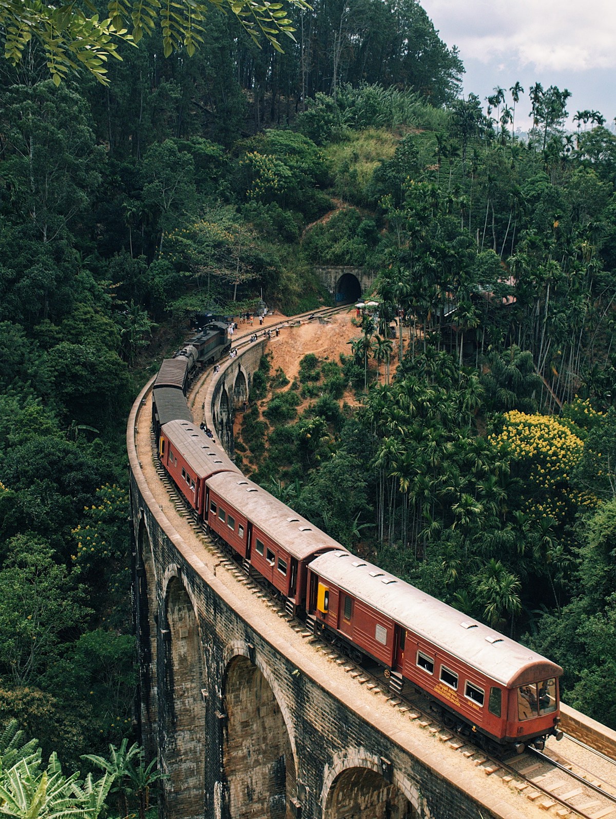 Red train crossing the Nine Arches Bridge viaduct surrounded by dense green forest in the Sri Lanka hill country